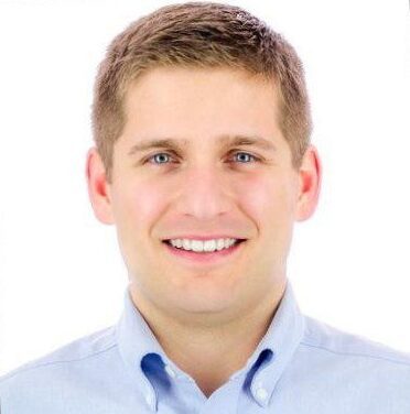 1617041843145 A man with short, light brown hair smiles while wearing a light blue button-up shirt. The background is plain white.